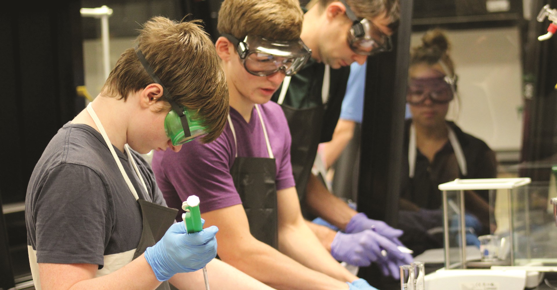 Image shows four students wearing safety goggles, gloves, and aprons participating in a chemistry lab experiment